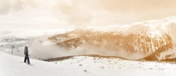 Panorama avec snowboarder assis avec snowboard dans les mains est assis sur un grand rocher sur la vue sur les montagnes. Bansko, Bulgarie — Photo