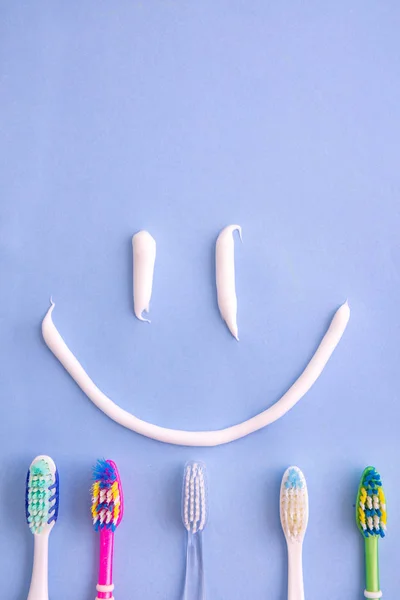 Pasta de dientes pequeña y varios cepillos de dientes como una sonrisa desde la vista superior en azul —  Fotos de Stock