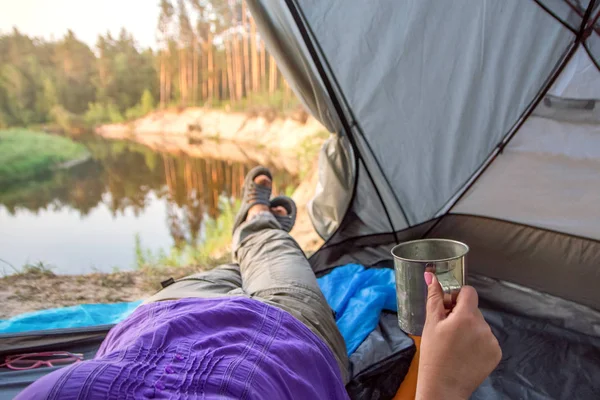 Fötter kvinna avkopplande njuter kaffe. Flod utsikt från tält Camping entré. Resor koncept äventyrs semestrar utomhus — Stockfoto