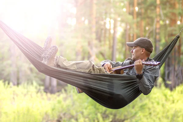 Retrato de un joven con sombrero tocando la guitarra y balanceándose en hamaca de fin de semana por la mañana en el campamento de descanso — Foto de Stock