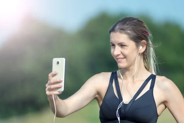 Chica sexy deportiva de fitness haciendo foto selfie en el teléfono inteligente durante el descanso de entrenamiento. Parque de la ciudad y cielo azul en el fondo —  Fotos de Stock
