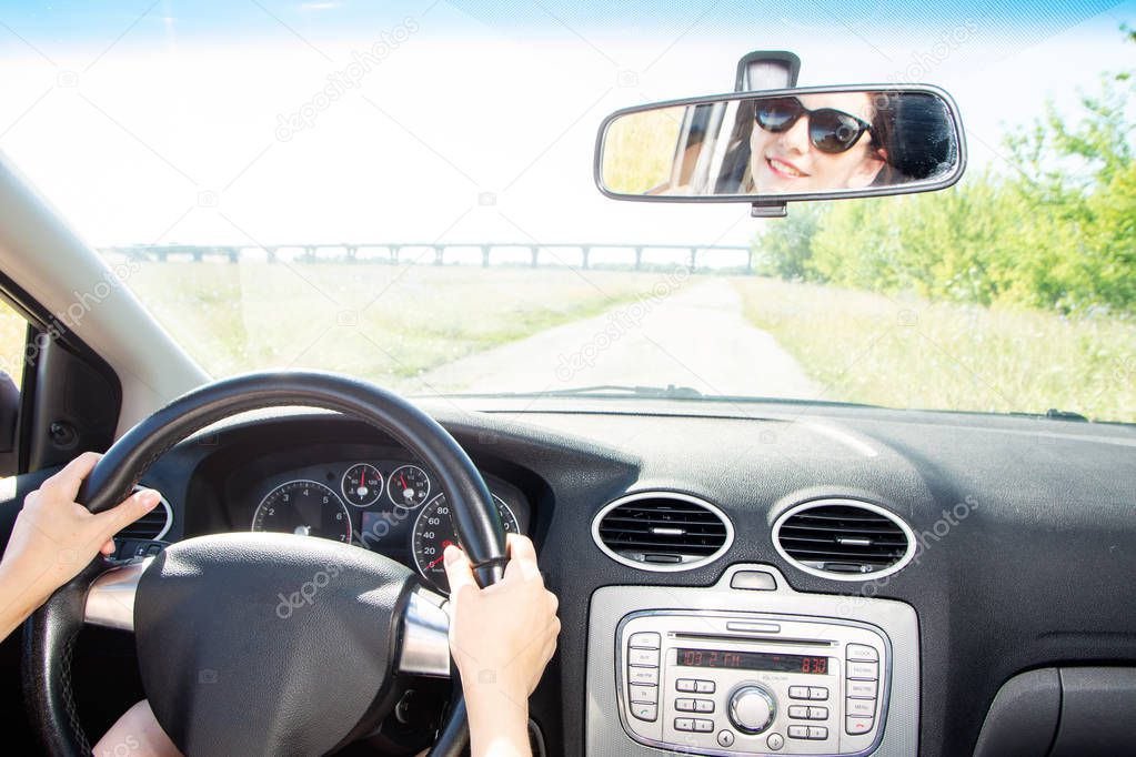 Young cheerful woman driving a car, rear view, reflexion of face in a mirror