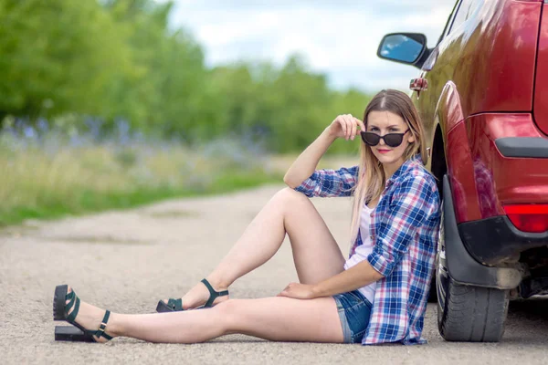 Widok z boku piękna dziewczyna siedzi w pobliżu samochodu na tle zielonego pola ze światłem słonecznym — Zdjęcie stockowe