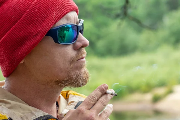 Hombre brutal en gafas de sol negro fumar un cigarrillo en el bosque verde, retrato hipster al aire libre —  Fotos de Stock