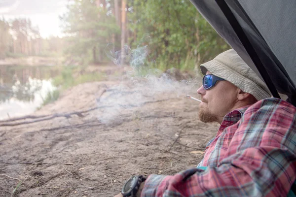 Man i en hatt och solglasögon som ligger i ett tält och röker en cigarett. koncept för resor och Camping — Stockfoto