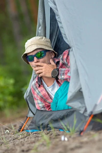 Hombre con sombrero y gafas de sol acostado en una tienda y fumando un cigarrillo. concepto de viaje y camping —  Fotos de Stock