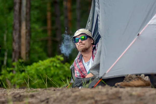 Man i en hatt och solglasögon som ligger i ett tält och röker en cigarett. koncept för resor och Camping — Stockfoto