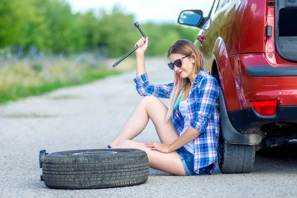 Hermosa chica con la llave del cilindro cambia la rueda en el coche roto. Neumáticos de recambio a un lado de la carretera . Fotos De Stock