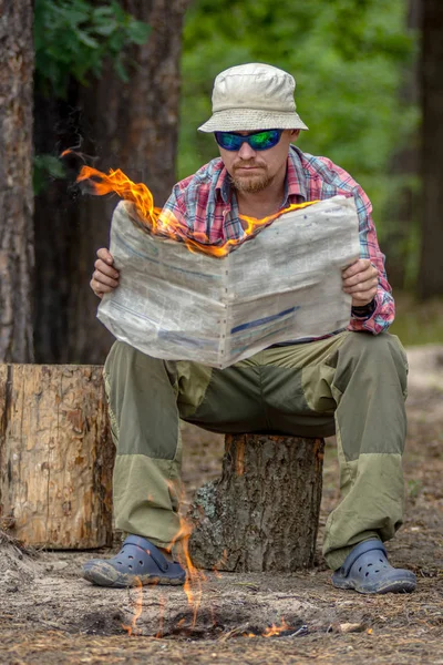 Turista sentado en el tocón y leyendo un periódico en llamas en el bosque cerca de la fogata. concepto noticias calientes — Foto de Stock