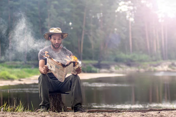 Turista sentado en el tocón y leyendo un periódico en llamas cerca del río en el bosque. concepto noticias calientes y supervivencia — Foto de Stock