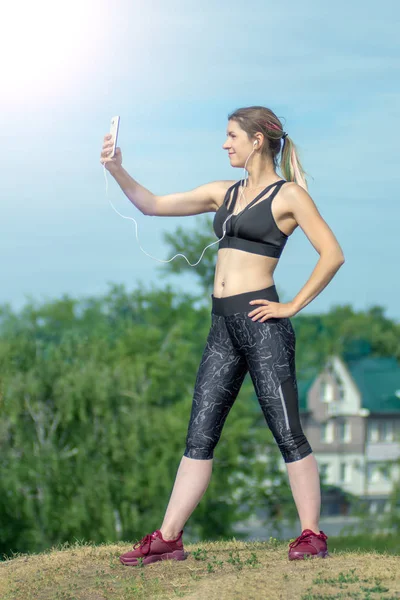 Chica sexy deportiva de fitness haciendo foto selfie en el teléfono inteligente durante el descanso de entrenamiento. Parque de la ciudad y cielo azul en el fondo —  Fotos de Stock