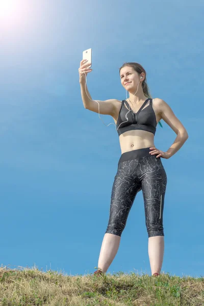 Chica sexy deportiva de fitness haciendo foto selfie en el teléfono inteligente durante el descanso de entrenamiento. Parque de la ciudad y cielo azul en el fondo —  Fotos de Stock