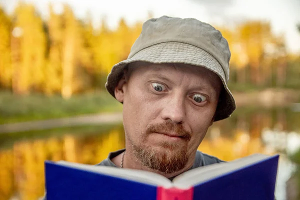 Funny tourist man in hat reading book at the beach near river and forest. concept survival book — Stock Photo, Image