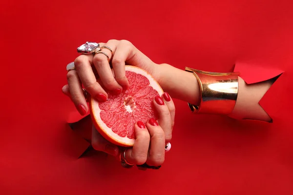 フルーツのマニキュア 赤い背景の穴から赤色の爪と女性の手は グレープ フルーツ果実を保持しています — ストック写真
