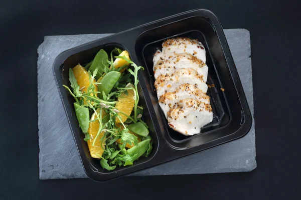 Balanced box diet, dinner dish. Poultry fillet on a salad with sugar peas and oranges on a black plate.