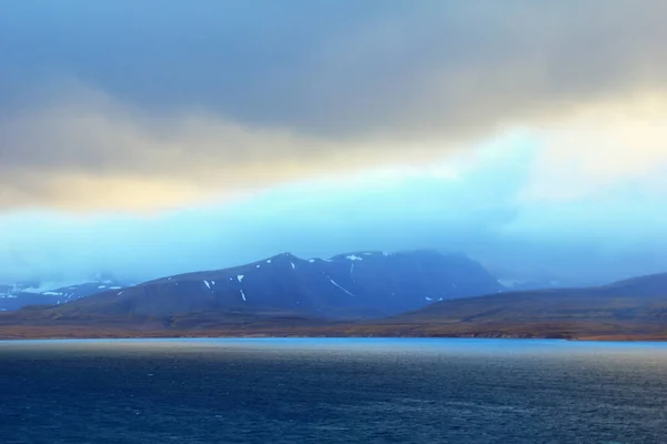 Güzel kutup manzarası - Barentsburg, Spitsbergen takımadaları (Svalbard Adası), Norveç, Grönland denizi yakınlarındaki dramatik bulutlu gökyüzünün arka planında kar ve mavi körfez ile kaplı siyah dağ