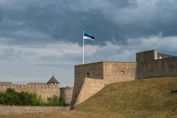 Bastião do castelo medieval Narva Herman. Estónia. Em primeiro plano está a bandeira da Estónia. No fundo é uma antiga fortaleza russa em Ivangorod. O rio Narva compartilha as fortalezas — Fotografia de Stock