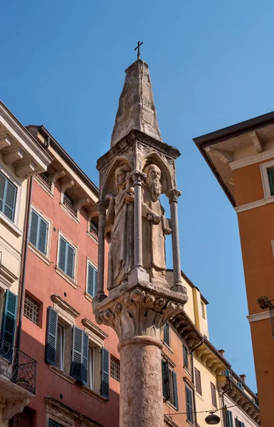Una antigua columna en la calle de Verona. Una pequeña columna gótica con esculturas de Nuestra Señora y de los Santos Cristóbal y Pedro el Mártir. La columna se encuentra entre las casas antiguas con persianas de madera — Foto de Stock