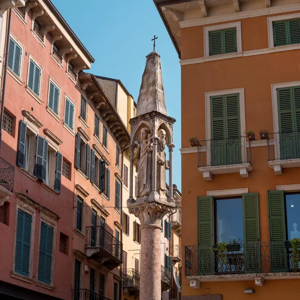 Una antigua columna en la calle de Verona. Una pequeña columna gótica con esculturas de Nuestra Señora y de los Santos Cristóbal y Pedro el Mártir. La columna se encuentra entre las casas antiguas con persianas de madera — Foto de Stock
