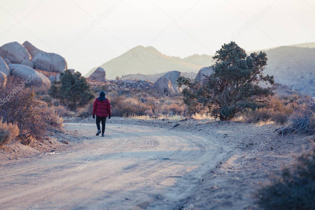 Man dressed in cold weather clothing walks along a dusty dirt road in the desert with huge boulders nearby
