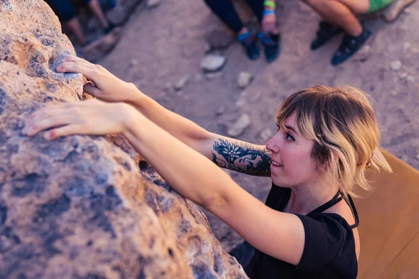 Thin blonde caucasian woman with arm tattoos struggles while rock climbing on boulders in the desert of California