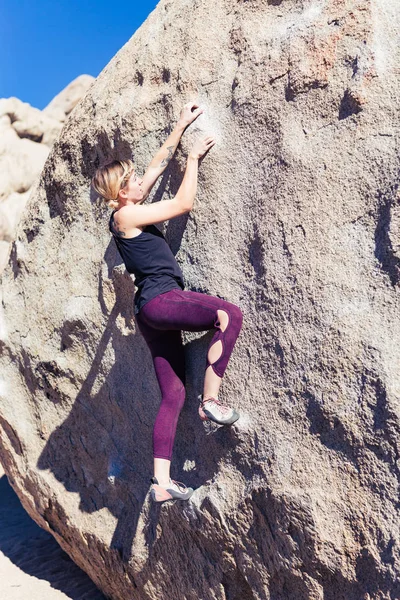 Thin blonde caucasian woman wearing black tank top rock climbing on boulders in the desert of California on a bright sunny day