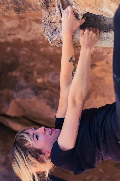 Thin blonde caucasian woman with arm tattoos hangs upside down while she rock climbs on boulders in the desert of California