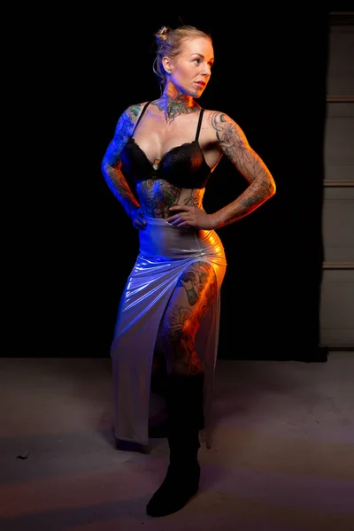 Muscled caucasian woman with tattoos and oiled skin wearing shiny skirt poses under colored lights