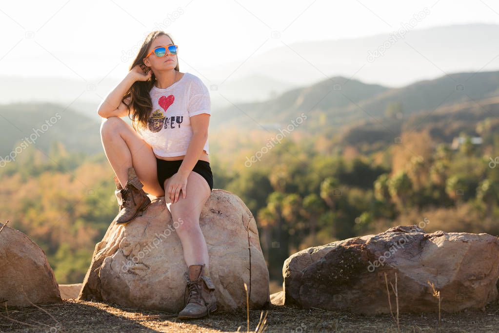 Young mixed race woman in white shirt and short shorts posing on a boulder near a forest destroyed by wildfire