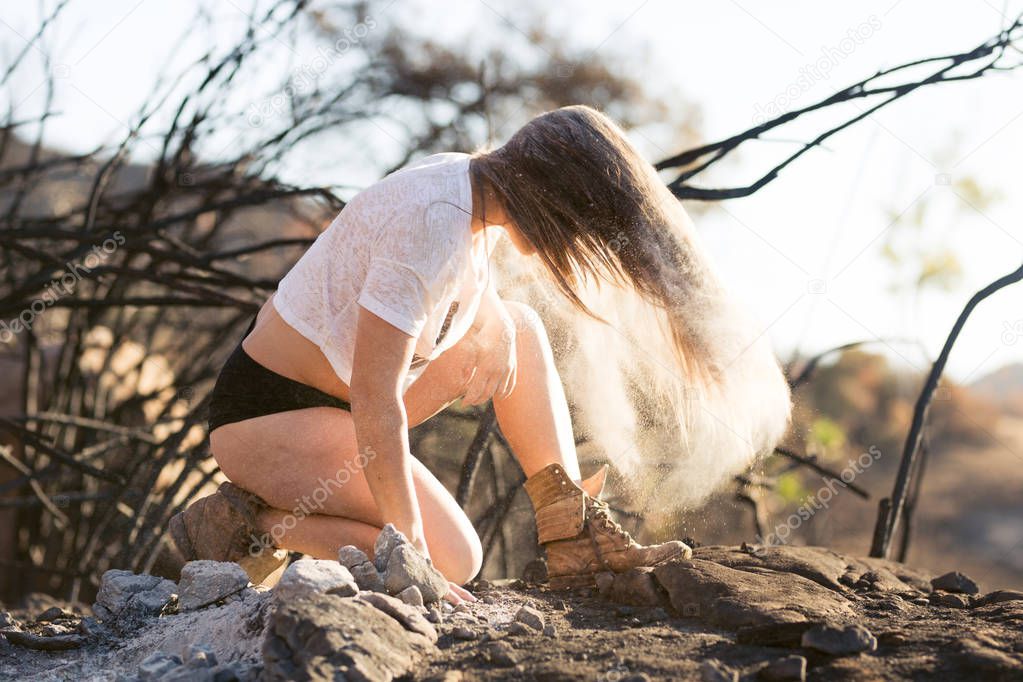 Young mixed race woman in white shirt and short shorts playing with hair near forest destroyed by wildfire while covered in ashes