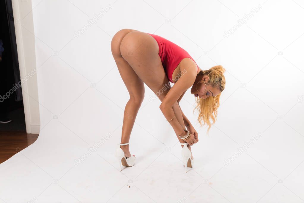 Fit young black  woman with long hair poses in a red one piece and white high heels against a white background