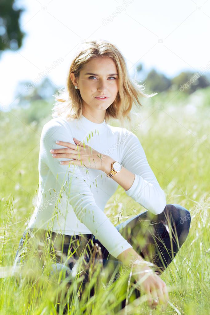 Attractive young blonde Caucasian woman in tight white top and denim posing outside in long grass