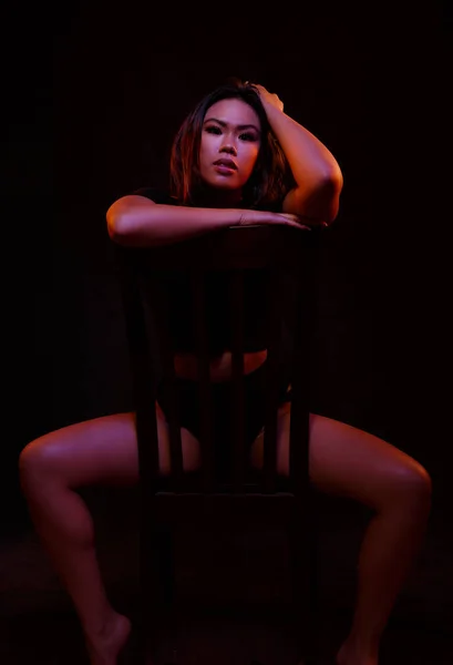 色のライトの下で椅子の上の黒のワンピース ポーズでフィットの若いアジア女性 — ストック写真