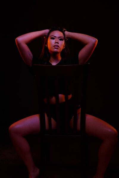 Fit young asian woman in black one piece poses on a chair under colored lights