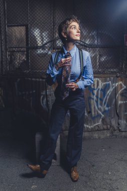 Takım elbise ve kravat giymiş queer kadın grungy endüstriyel bir yerde pozlar