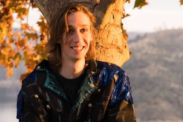 Young man with long hair poses against tree at sunset above scenic lake