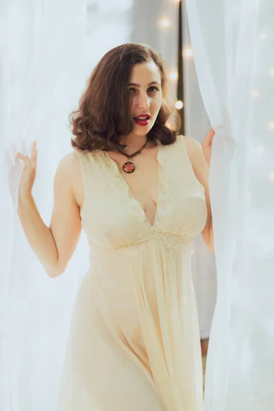 Young woman in retro pin up style and white dress poses against a white curtain backdrop