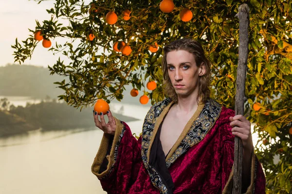 Young caucasian man with long hair and royal costume clothing lounges under an orange tree