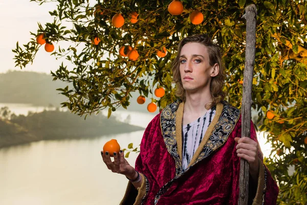 Young caucasian man with long hair and royal costume clothing lounges under an orange tree