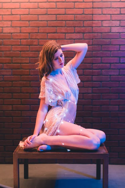 Young caucasian woman in sheer dress poses with string lights against a brick wall