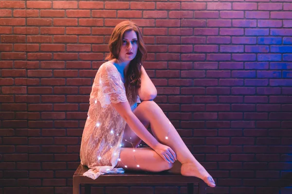 Young caucasian woman in sheer dress poses with string lights against a brick wall