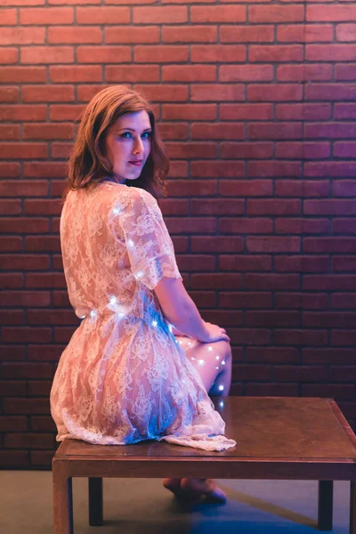Young caucasian woman in sheer dress poses with string lights against a brick wall