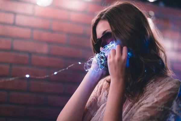 Young caucasian woman in sheer dress poses with string lights against a brick wall