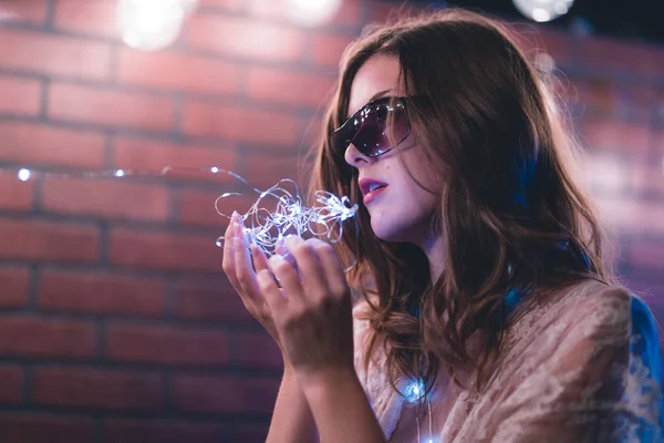 Young caucasian woman in sheer dress poses with string lights against a brick wall