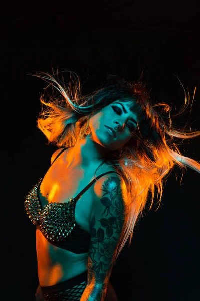 Alternative model with bangs and colored hair poses under teal and orange light wearing a fishnet top