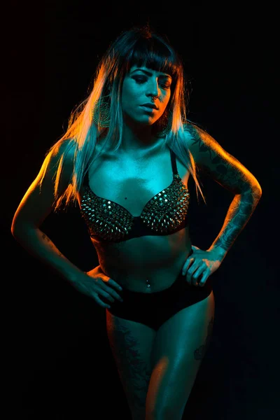 Alternative model with bangs and colored hair poses under teal and orange light wearing a fishnet top