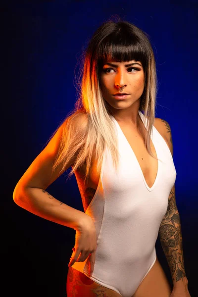 Alternative model with bangs and colored hair poses under blue and orange light wearing a white leotard