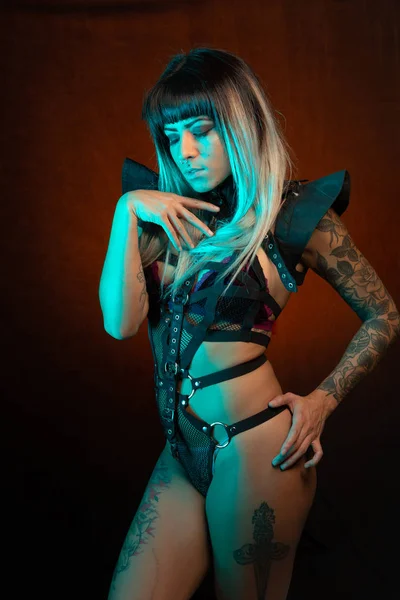 Alternative model with bangs and colored hair poses under blue light wearing a leather outfit