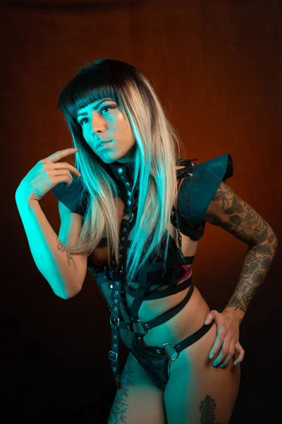 Alternative model with bangs and colored hair poses under blue light wearing a leather outfit