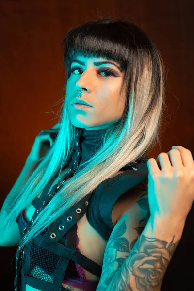 Alternative model with bangs and colored hair poses under blue light wearing a leather outfit